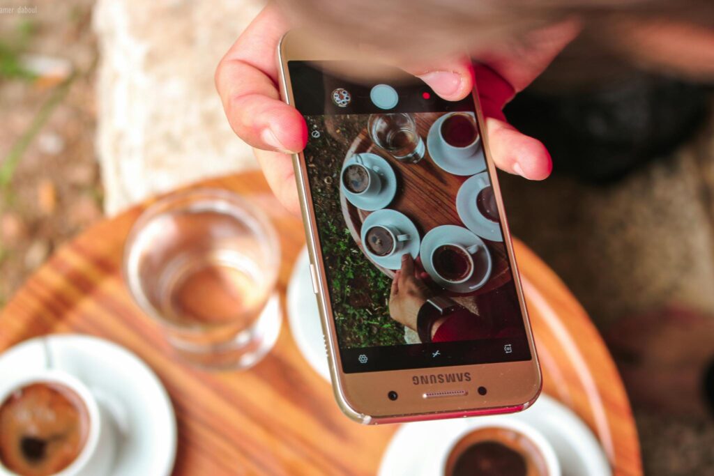 A hand holding a smartphone taking a photo of coffee cups on a wooden table outdoors.